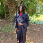 Joe Johnson in a grove of palm trees in a graduation gown holding a grad cap